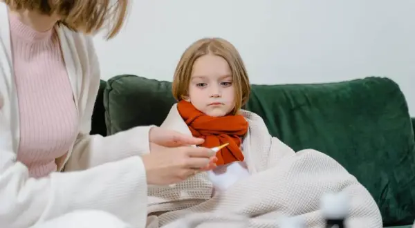 Smutna dziewczynka owinięta kocem, z szalikiem na szyi, czeka na powrót do zdrowia po grypie. Mama mierzy jej temperaturę.