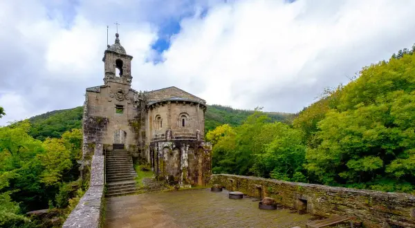Antiguo monasterio de San Juan de Caaveiro, con su torre campanario y ábside románico, rodeado de exuberante vegetación verde.