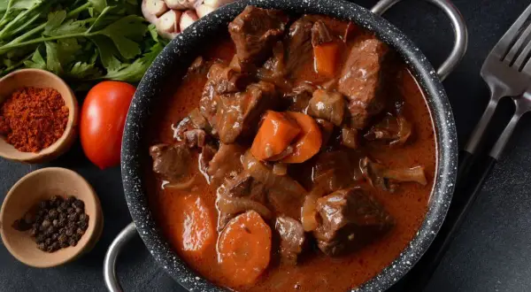 Gulasz z sarny w głębokim sosie, z marchewką i cebulą, obok przypraw i pomidorów.