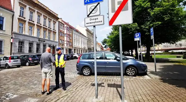 Policjant rozmawia z mężczyzną na brukowanej ulicy. Znaki drogowe wskazują strefę zamieszkania i koniec strefy parkowania.