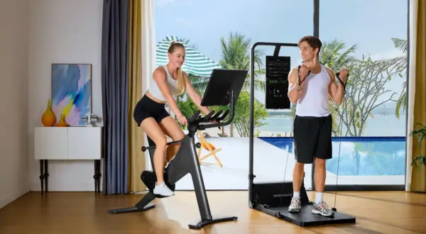 Ein Paar macht Ausdauertraining zuhause: Sie auf dem Fahrrad, er mit Widerstandsbändern. Der Blick geht auf ein tropisches Paradies.