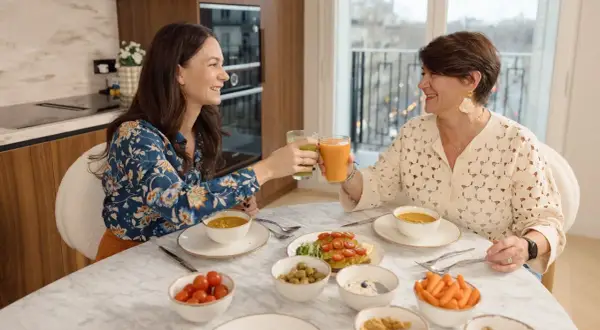 Deux femmes trinquant avec des jus verts et orangés, entourées d'un repas sain. Un exemple de comment ne pas grossir à la ménopause.