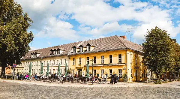 Gemütliche Straßencafés beleben die sonnige Rheinsberg Altstadt. Menschen sitzen unter grünen Sonnenschirmen vor gelben Fachwerkhäusern.