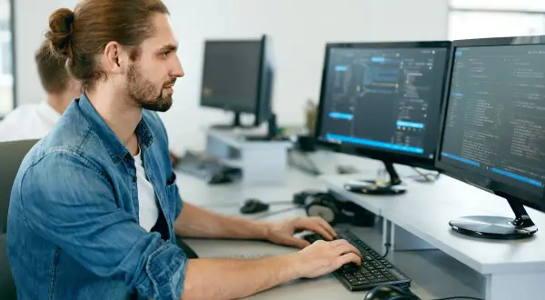 Młodszy programista z kitką na głowie pracuje przy dwóch monitorach wyświetlających kod.