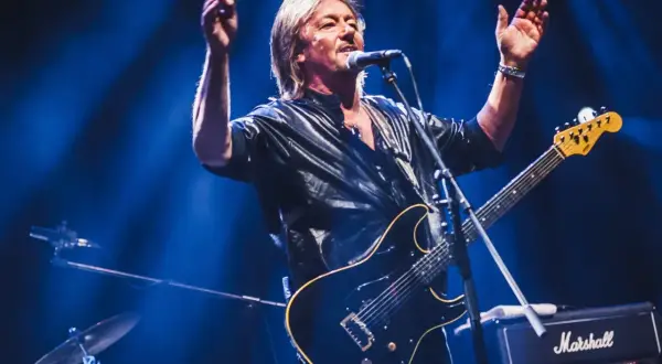 Chris Norman, lider zespołu Smokie, śpiewa i gra na gitarze na scenie, oświetlony niebieskim światłem.