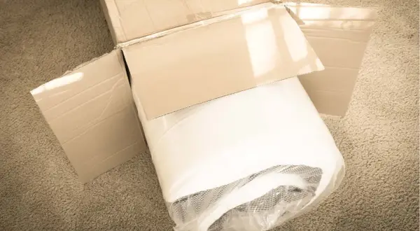 A rolled-up mattress, still in its plastic wrap, sits in an open cardboard box on a carpet. It looks like this left mattress in box too long.