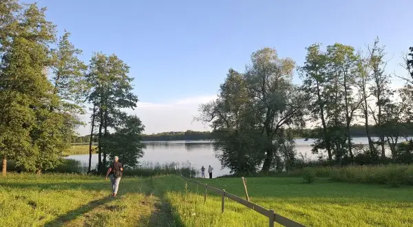 Piękny litewski krajobraz nad jeziorem. Osoba idzie ścieżką przez zieloną łąkę w stronę wody, gdzie stoją inni ludzie.