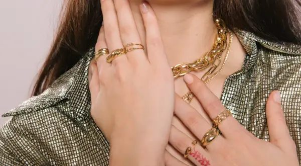 Manos adornadas con varios anillos dorados y un collar grueso, mostrando cómo llevar anillos de manera elegante.