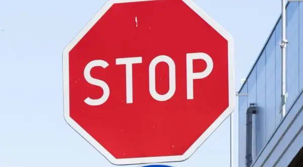 Znak b20 STOP. Czerwony ośmiokąt z białym napisem STOP. W tle błękitne niebo i fragment budynku.