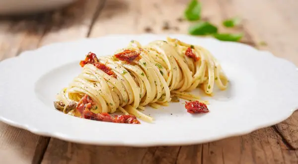 Un piatto di linguine condite con pomodori secchi, capperi e prezzemolo, su un piatto bianco con sfondo in legno.