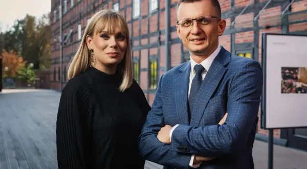 Para adwokatów, kobieta w czarnej bluzce i mężczyzna w niebieskiej marynarce, stoją przed ceglanym budynkiem.