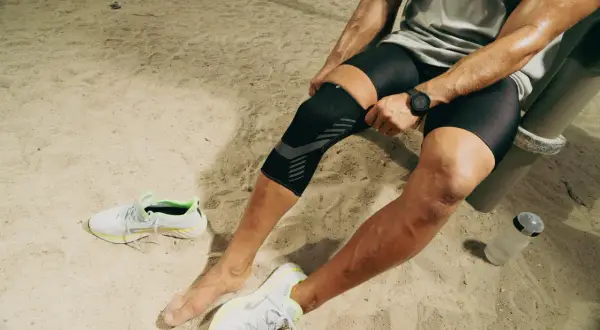 Un athlète ajuste sa genouillère TFL Decathlon sur un terrain sablonneux, prêt pour son entraînement.