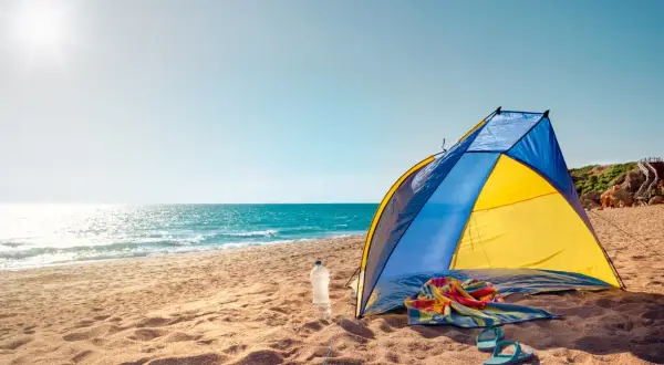 Kolorowy namiot na plaży, obok butelka wody, ręcznik i klapki. Słońce odbija się w morzu.