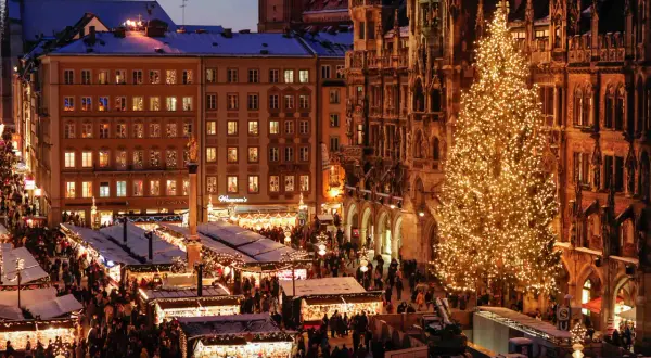 Beleuchteter Weihnachtsbaum und geschäftiger Weihnachtsmarkt bis Silvester, mit Buden und Menschen vor historischen Gebäuden.