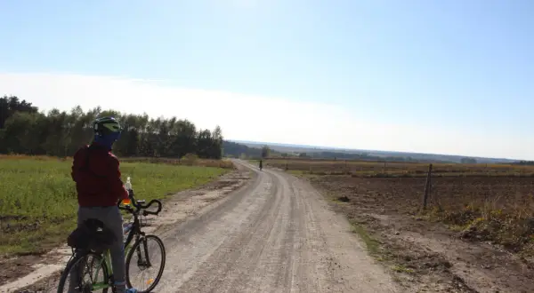 Rowerzysta na szutrowej drodze, która wydaje się być częścią najdłuższej trasy rowerowej w Polsce. Słońce świeci na błękitnym niebie.