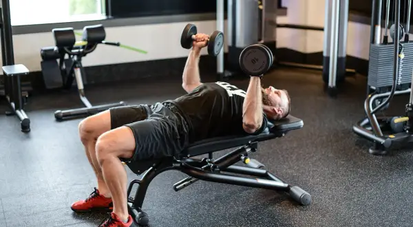 Mężczyzna wykonuje ćwiczenie dumbbell press na ławce w siłowni, podnosząc ciężarki nad klatką piersiową.