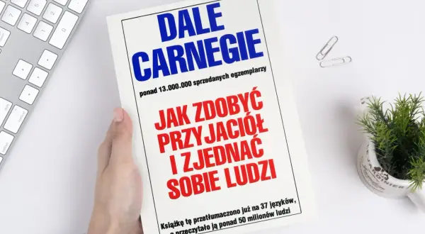 Książka "Jak zdobyć przyjaciół i zjednać sobie ludzi" autorstwa Dale'a Carnegie, trzymana w dłoni, leży obok klawiatury i rośliny.