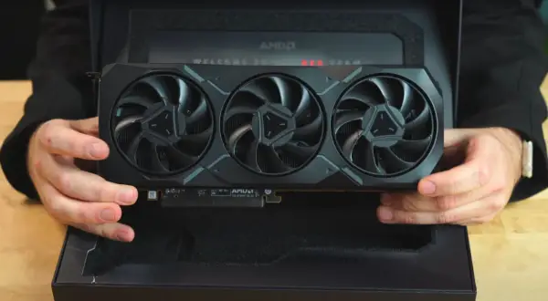 Ręce trzymają kartę graficzną Radeon RX 7900 XTX z trzema wentylatorami, wyjętą z czarnego pudełka z napisem "WELCOME TO RED TEAM".