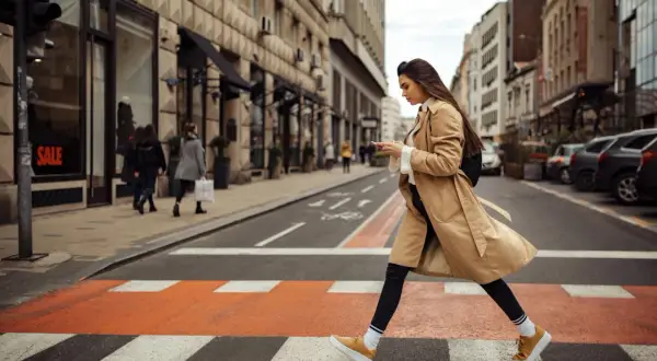 Kobieta w beżowym płaszczu i żółtych sneakersach przechodzi przez pasy. Idealne buty do trencza na miejski spacer.
