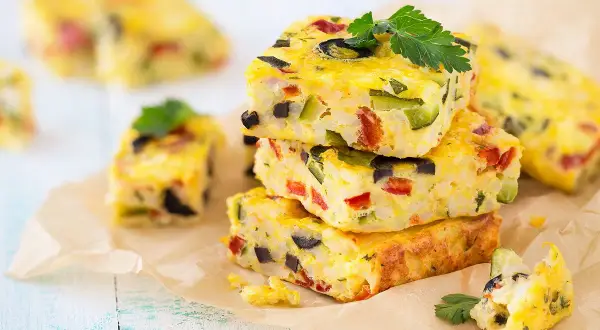 Carrés de frittata colorés, une recette avec des oeufs, garnie de légumes et d'olives, sur du papier sulfurisé.