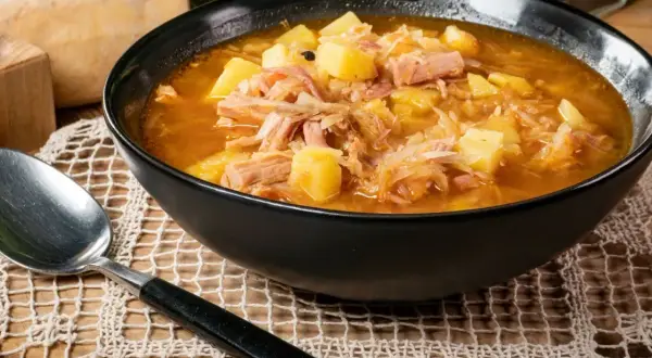 Parzybroda, sycąca zupa z kapusty kiszonej, ziemniaków i mięsa, podana w czarnej misce, obok leży łyżka.