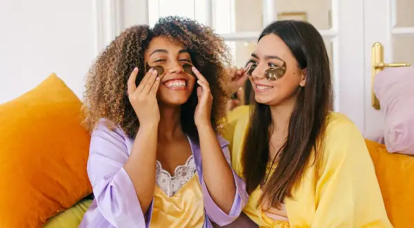 Due amiche sorridono mentre applicano patch per gli occhi, un essenziale skincare step, indossando vestaglie colorate.