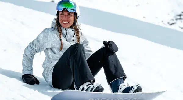 Uśmiechnięta snowboardzistka w kasku i goglach siedzi na śniegu obok deski. Jej pokrowiec na deskę snowboardową jest gotowy do drogi.