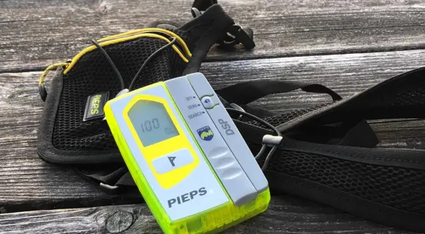 Ein Pieps lawinen sender mit Display und Suchfunktion, daneben eine schwarze Tasche mit gelben Akzenten, liegt auf einer Holzoberfläche.