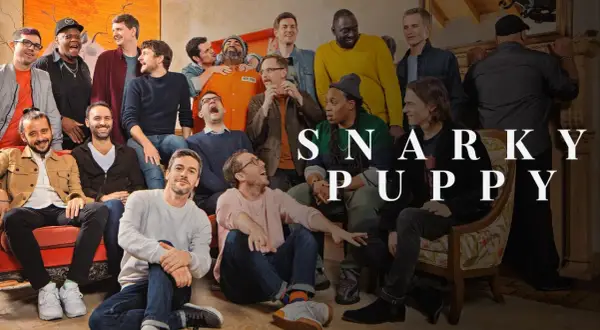 Grupa ludzi, w tym mężczyzna w pomarańczowym kombinezonie, pozuje do zdjęcia z napisem "SNARKY PUPPY".