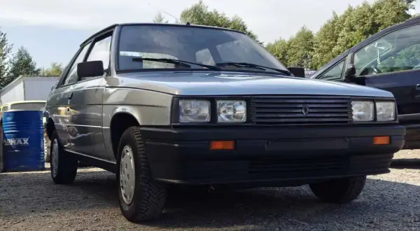Srebrny Renault 11 z lat 80. stoi na żwirowym podjeździe, obok niebieskiego beczki i innego samochodu.