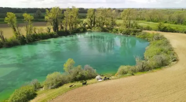 Idyllischer Paylake in Frankreich mit türkisfarbenem Wasser, umgeben von grüner Vegetation und Feldern. Angler haben ihr Lager aufgeschlagen.