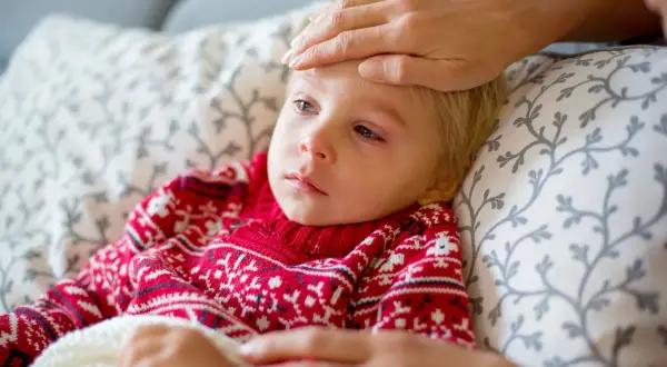 Małe dziecko w czerwonym sweterku z wzorem płatków śniegu leży chore, z gorączką. Ręka opiekuna sprawdza temperaturę. To typowy obrazek, gdy dopada nas wirus.