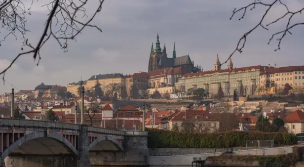 Praga nietypowe atrakcje: widok na Hradczany i Most Karola przez Wełtawę, z gałęziami drzew w pierwszym planie.