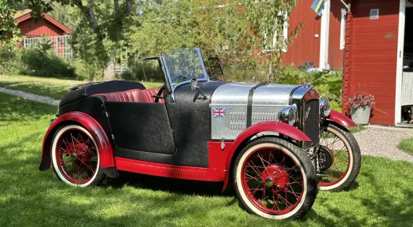 En klassisk Austin bilmodell i svart och rött med brittisk flagga, parkerad på en gräsmatta framför ett rött hus med svensk flagga.