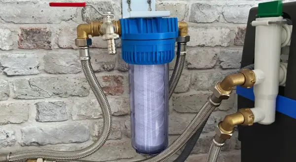 Filtre à eau avec cartouche visible, raccordé à un adoucisseur qui coule en permanence. Tuyaux flexibles et robinet rouge.