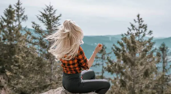 Frau mit wehendem blonden Haar sitzt auf einem Felsen im bayerischen Wald und genießt die Aussicht.