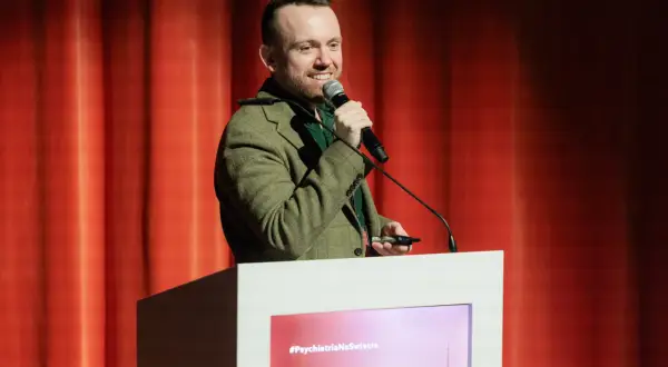 Mężczyzna w tweedowej marynarce przemawia na tle czerwonej kurtyny. Mówi o psychiatrii i klozapinie, trzymając mikrofon.
