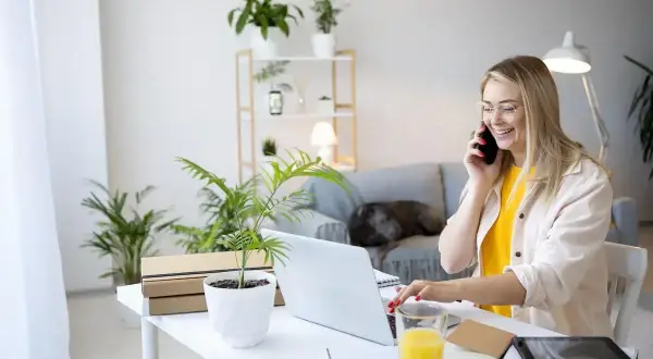 Uśmiechnięta kobieta rozmawia przez telefon przy biurku z laptopem i roślinami.