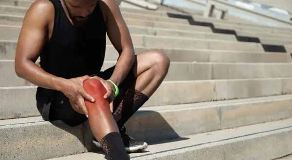 Sportler mit schmerzhaft gerötetem Knie auf Treppen. Er hofft, seine Gelenkschmiere aufbauen zu können, um bald wieder fit zu sein.