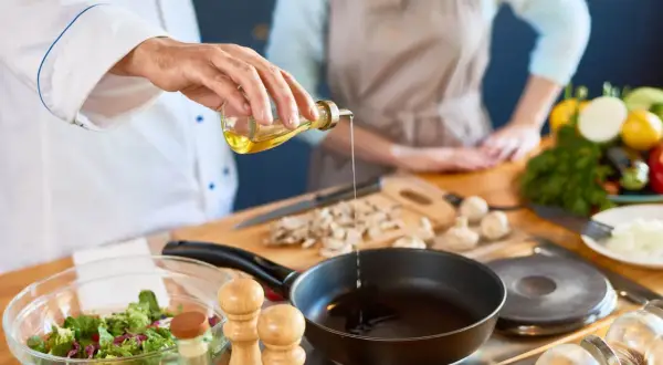 Szef kuchni nalewa oliwę z oliwek do gorącej patelni, przygotowując się do smażenia frytek. W tle widać warzywa i składniki.