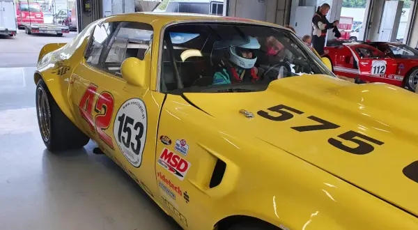 Gul Firebird 69 med nummer 153 och 575 redo för race. En förare i hjälm sitter bakom ratten.
