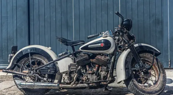 Stary motocykl Harley-Davidson w kolorze białym i czarnym, stojący na bruku przed niebieską, drewnianą ścianą.