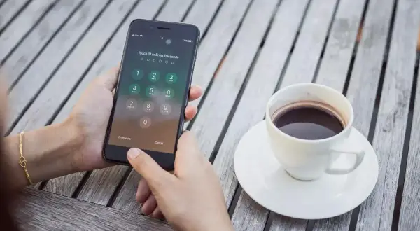 Zablokowany iPhone z klawiaturą numeryczną i ikoną Touch ID. Obok filiżanka kawy na drewnianym stole.
