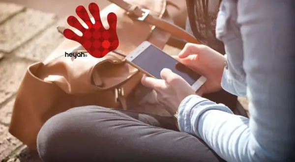 Kobieta z telefonem w dłoniach, obok brązowa torba i czerwona dłoń z logo heyah.