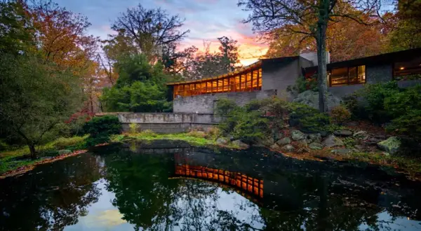 The last Wright house, a modern masterpiece, reflects warmly in a tranquil pond amidst autumn foliage.