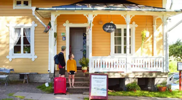 Två personer med resväska anländer till Hajstorp Vandrarhem. En skylt välkomnar besökare.