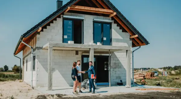 Para młoda uśmiecha się do doradcy, który wręcza im dokumenty. W tle widać dom w budowie, symbolizujący ich marzenie o własnym lokum, które zrealizują dzięki kredytowi na budowę domu.