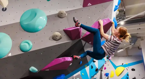 Frau klettert an einer bunten Wand in einer Boulderhalle in Hamburg. Sie hält sich an einem pinken Griff fest.