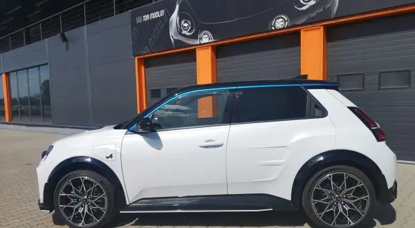 Biały Alpine A290 z niebieskimi akcentami stoi przed budynkiem z pomarańczowymi drzwiami garażowymi.