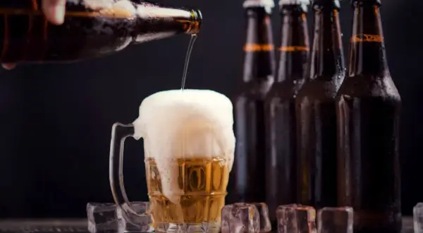 Una mano versa birra in un bicchiere traboccante di schiuma. Sullo sfondo, bottiglie di ibu birra e cubetti di ghiaccio.